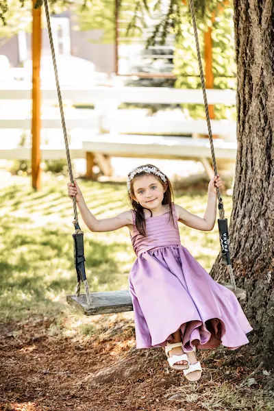 Flower girl in muave dress smiling while sitting on a wooden tree swing